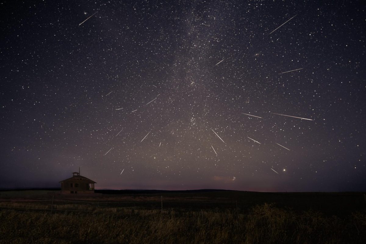 Image of night sky with multiple meteor streaks throughout. Bottom third depicts a field with an abandoned school towards the left side.