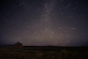 Image of night sky with multiple meteor streaks throughout. Bottom third depicts a field with an abandoned school towards the left side.